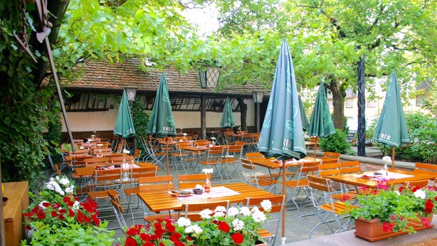 An outdoor café with wooden tables and chairs, green umbrellas, and potted plants. - Hotel Petzengarten (Nuremberg)