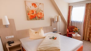 A hotel room with a neatly made bed, a bedside table with a lamp, a telephone, and a power outlet. There is a painting of flowers on the wall, a chair with a floral cushion, and a window with curtains. - Hotel Petzengarten (Nuremberg)