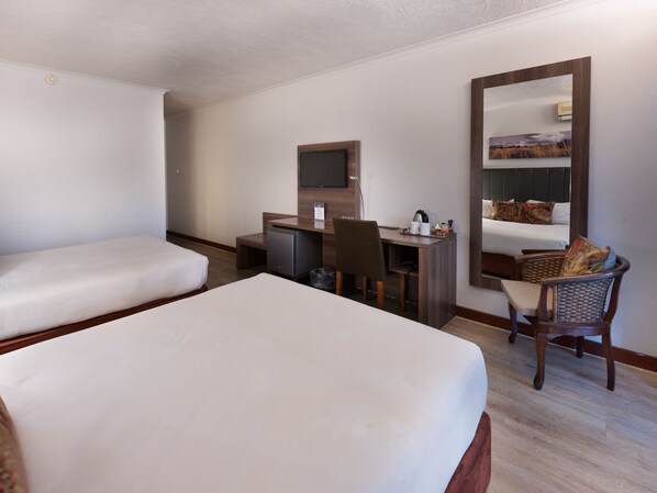 A hotel room with two single beds, a desk with a chair, a small refrigerator, a mirror, and a window.