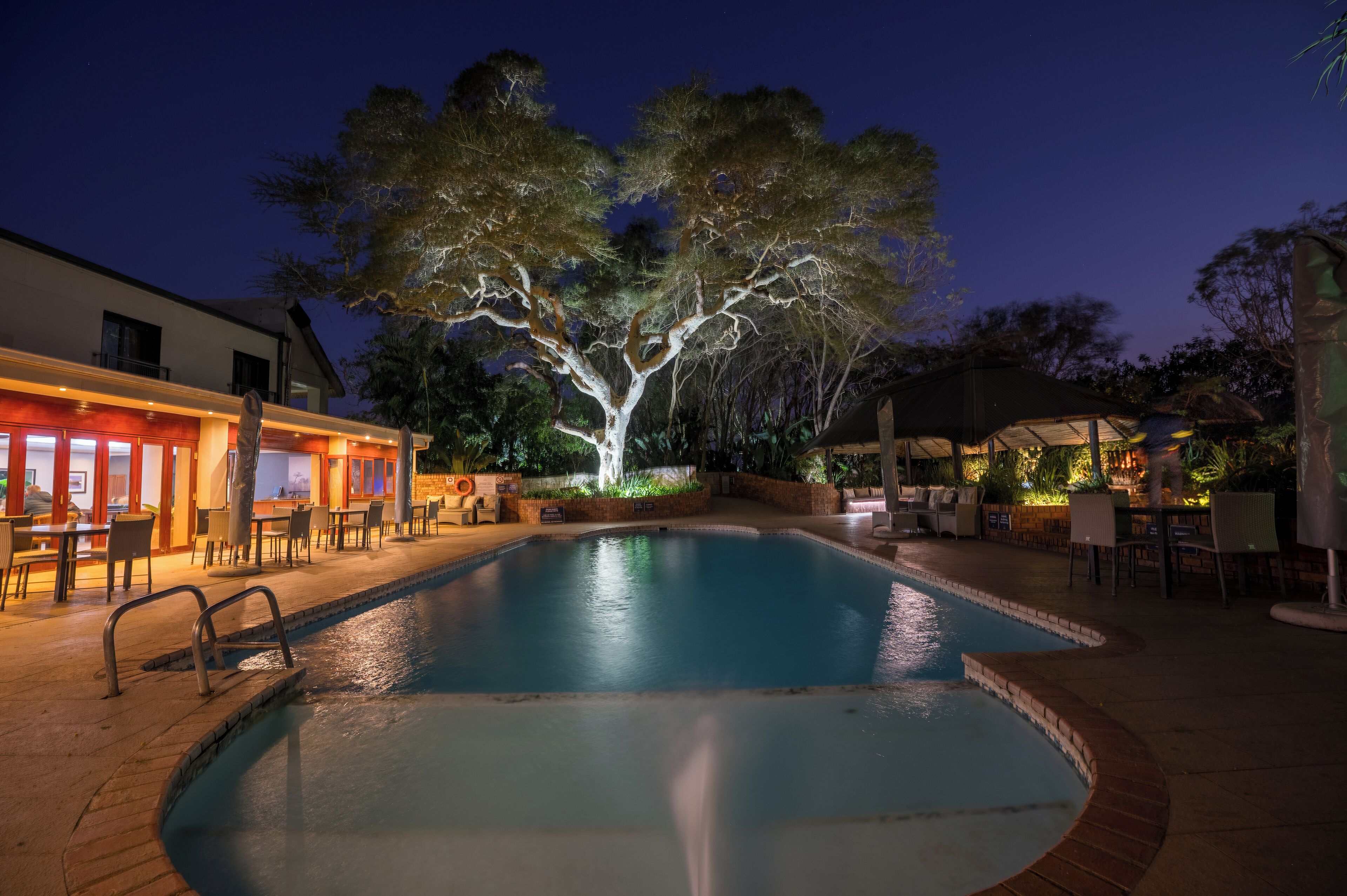 A swimming pool with a tree in the center, surrounded by outdoor seating and a building with a bar area.