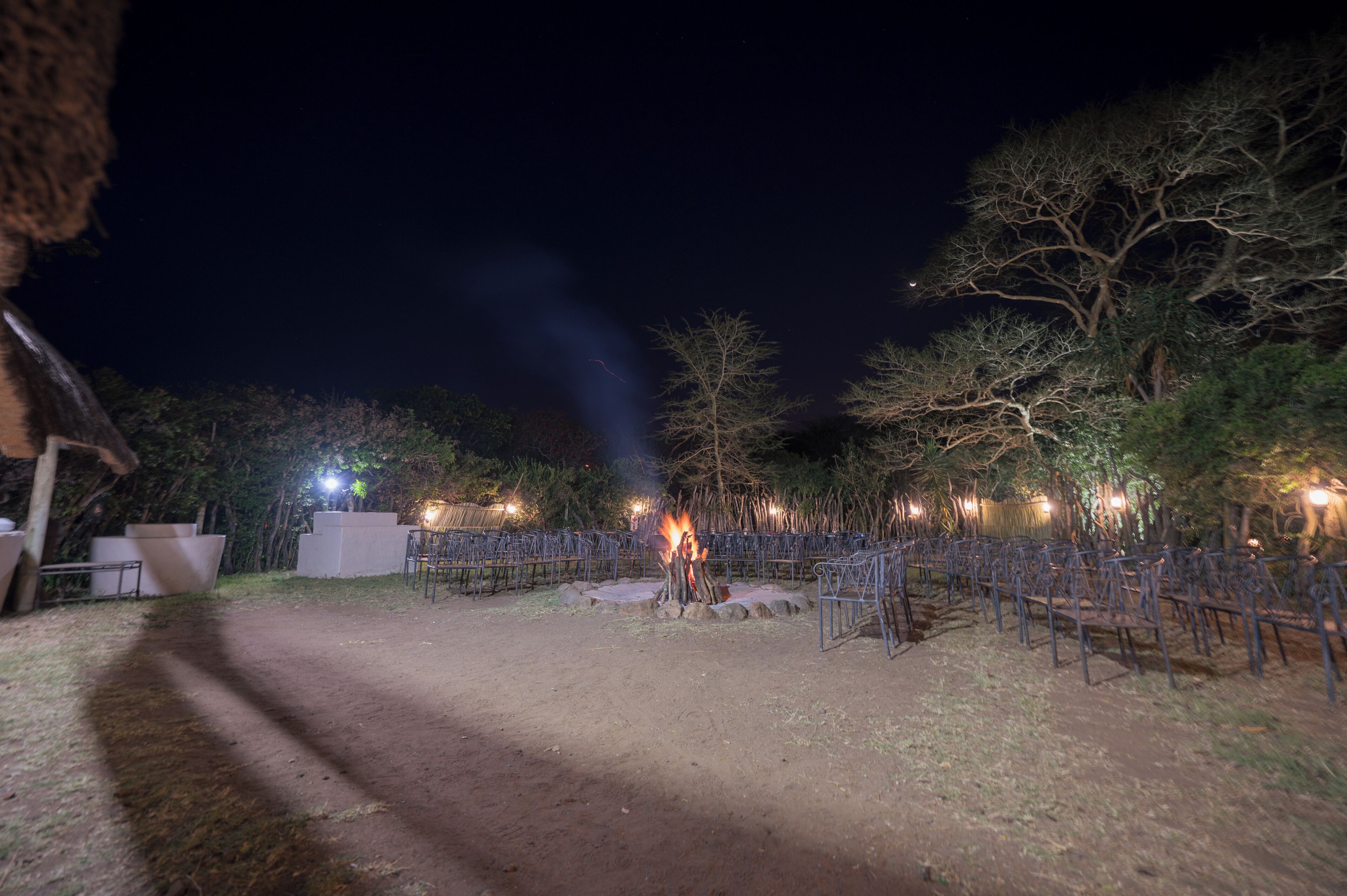 A nighttime outdoor area with a fire, seating arrangements, and trees.