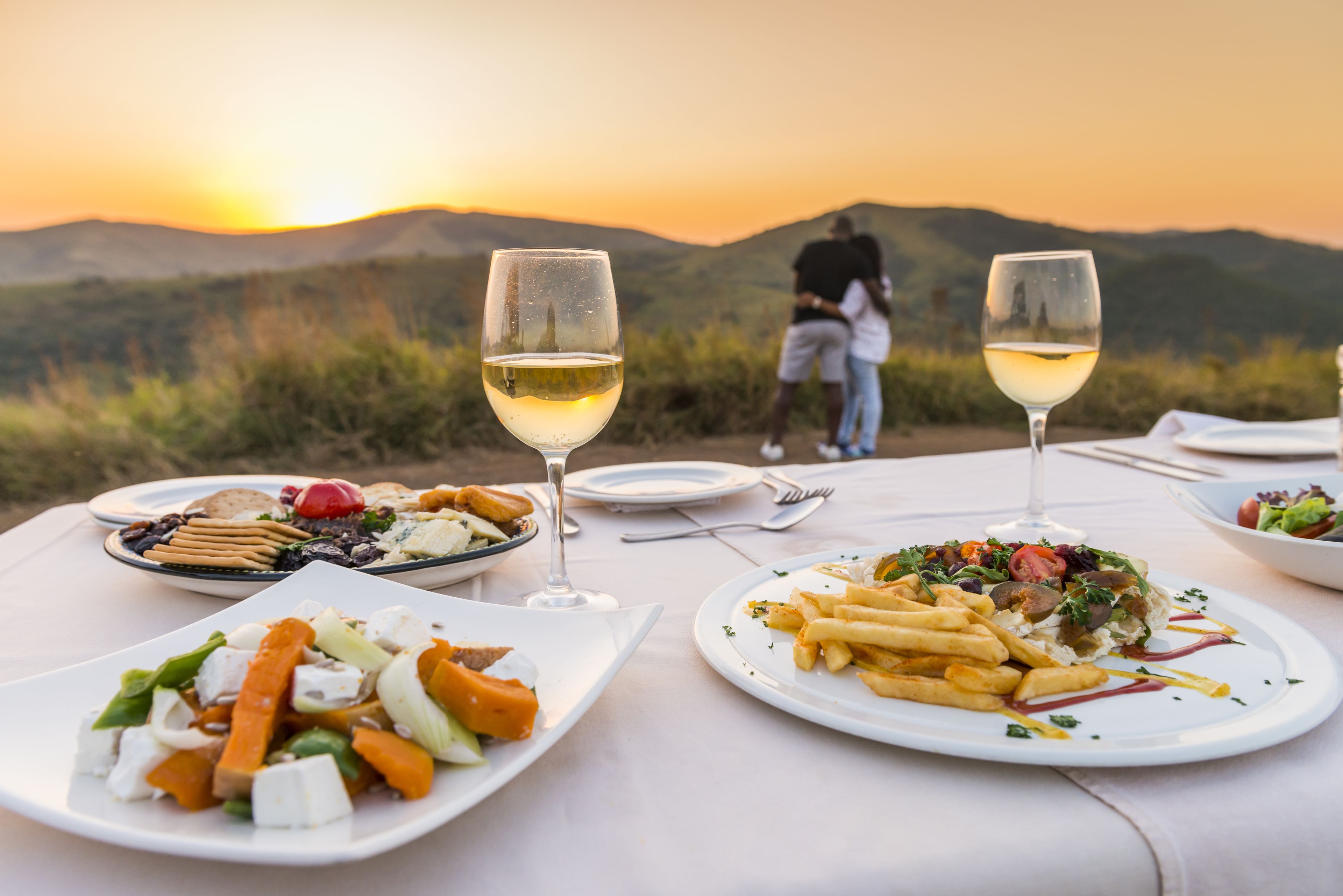 A table set for a meal with plates of food and glasses of white wine, overlooking a mountainous landscape.