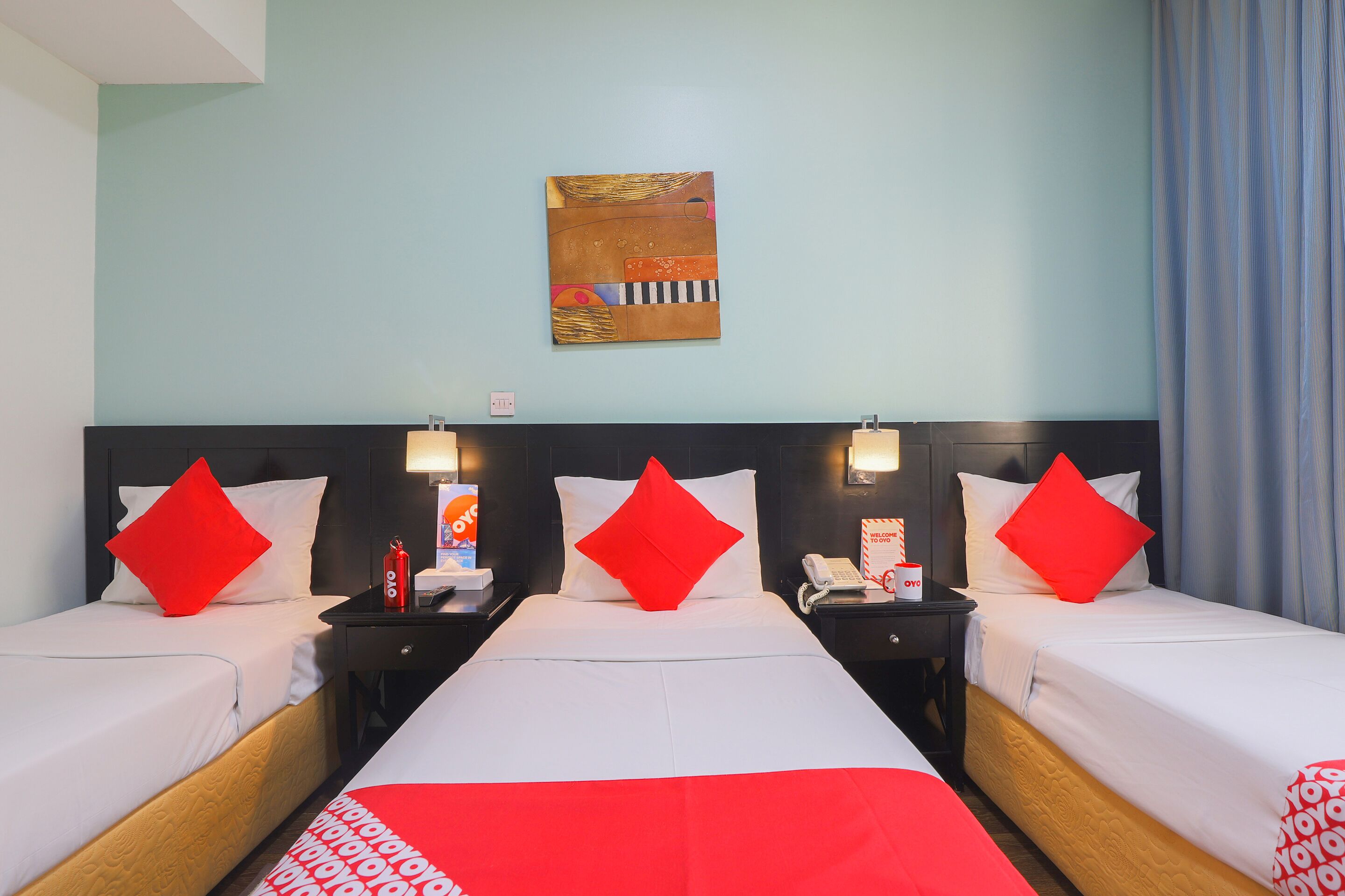 A hotel room with two beds, each with white bedding and red pillows. There is a painting on the wall above the beds, a nightstand with a lamp and a phone, and a window with curtains.