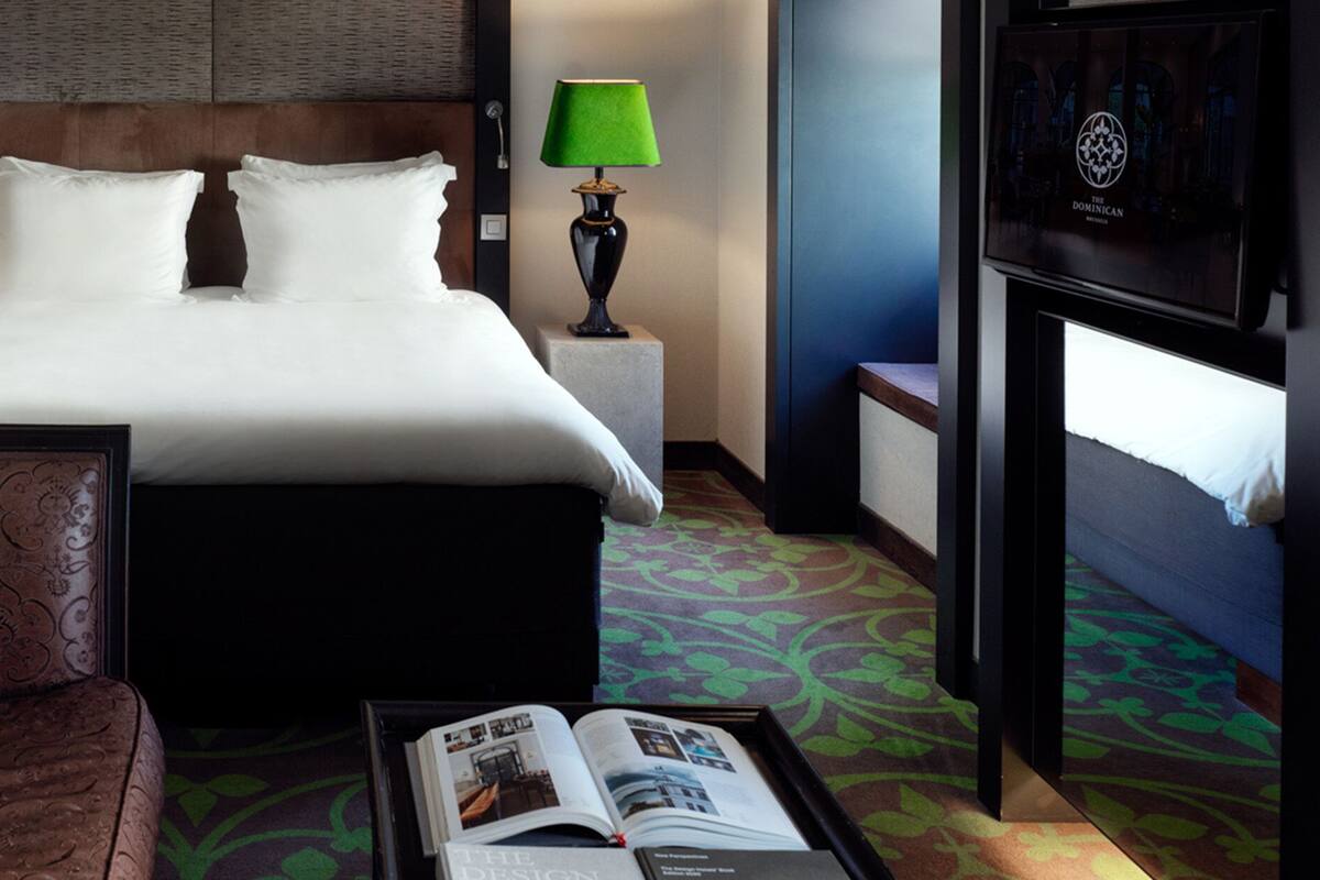 Une chambre d’hôtel avec un lit, une table de chevet avec une lampe, un tableau encadré au mur et un magazine sur le sol.