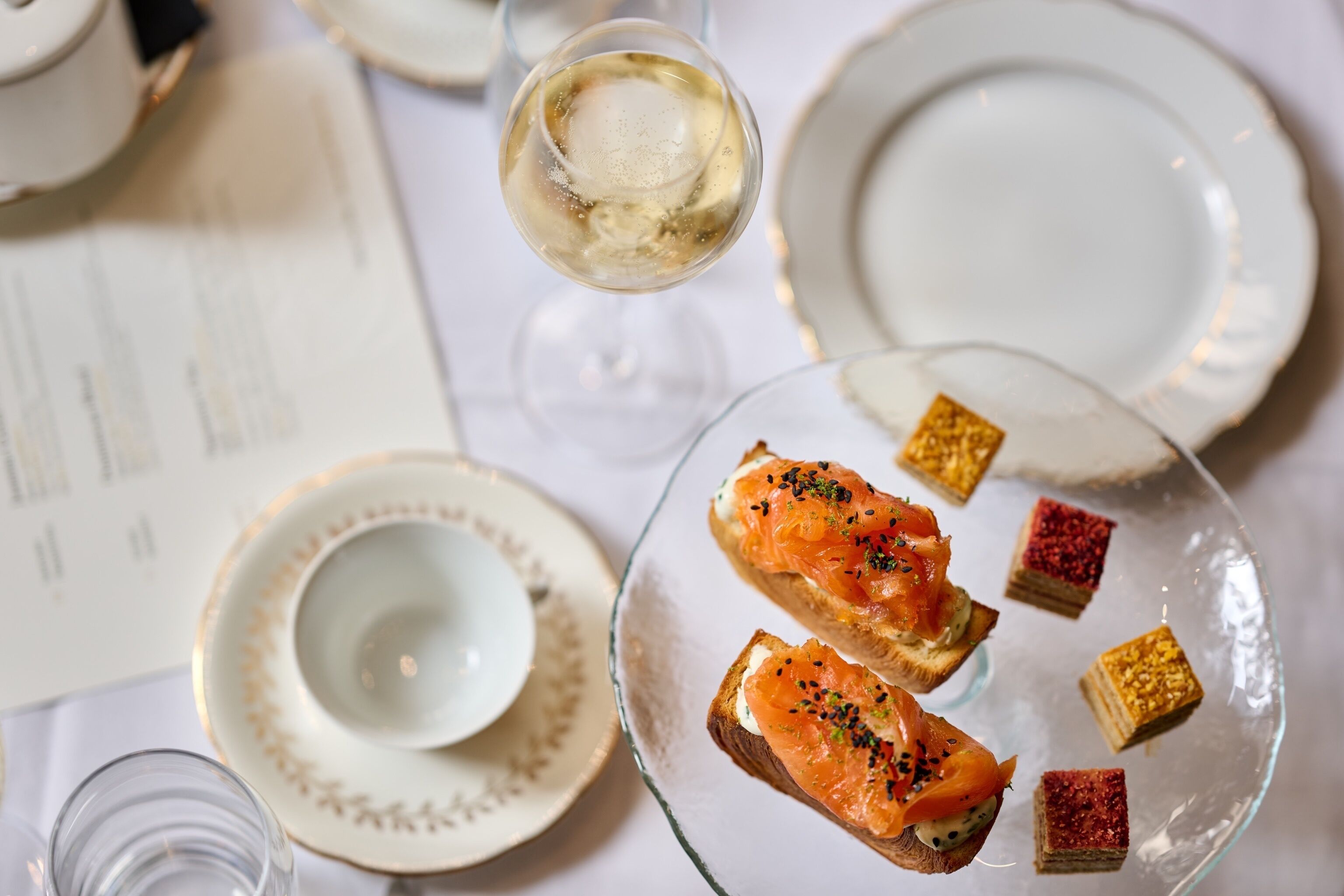 Une table avec un verre de vin blanc, une assiette de toasts garnis de saumon fumé et de petits desserts carrés.