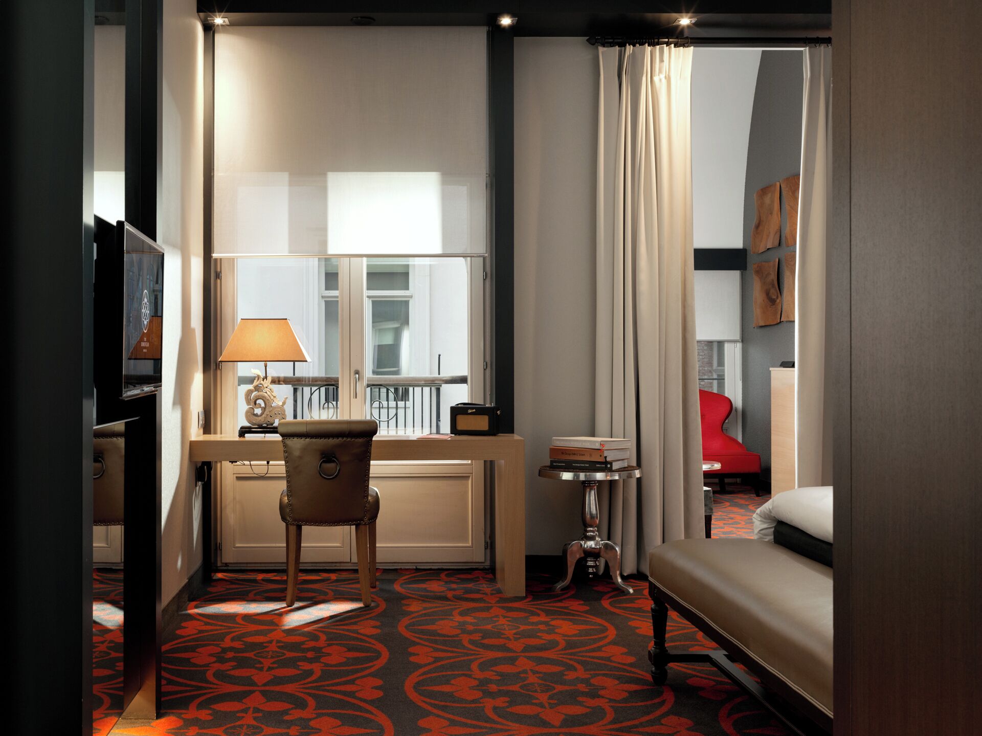 A hotel room with a bed, a desk with a lamp, a chair, a small table, and a window with curtains.