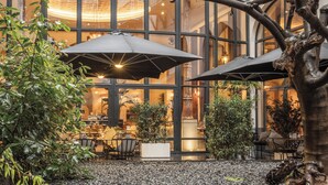 An outdoor seating area with umbrellas, tables, and chairs, surrounded by plants and a large glass building in the background. - The Dominican, Brussels, a Member of Design Hotels (Brussels)