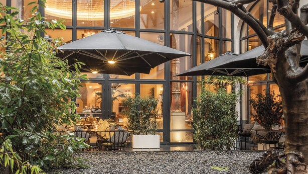 An outdoor seating area with umbrellas, tables, and chairs, surrounded by plants and a large glass building in the background. - The Dominican, Brussels, a Member of Design Hotels (Brussels)