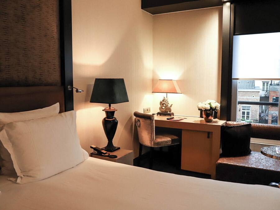 Habitación de hotel con cama, escritorio con lámpara, silla, mesita y ventana con vista a edificios.