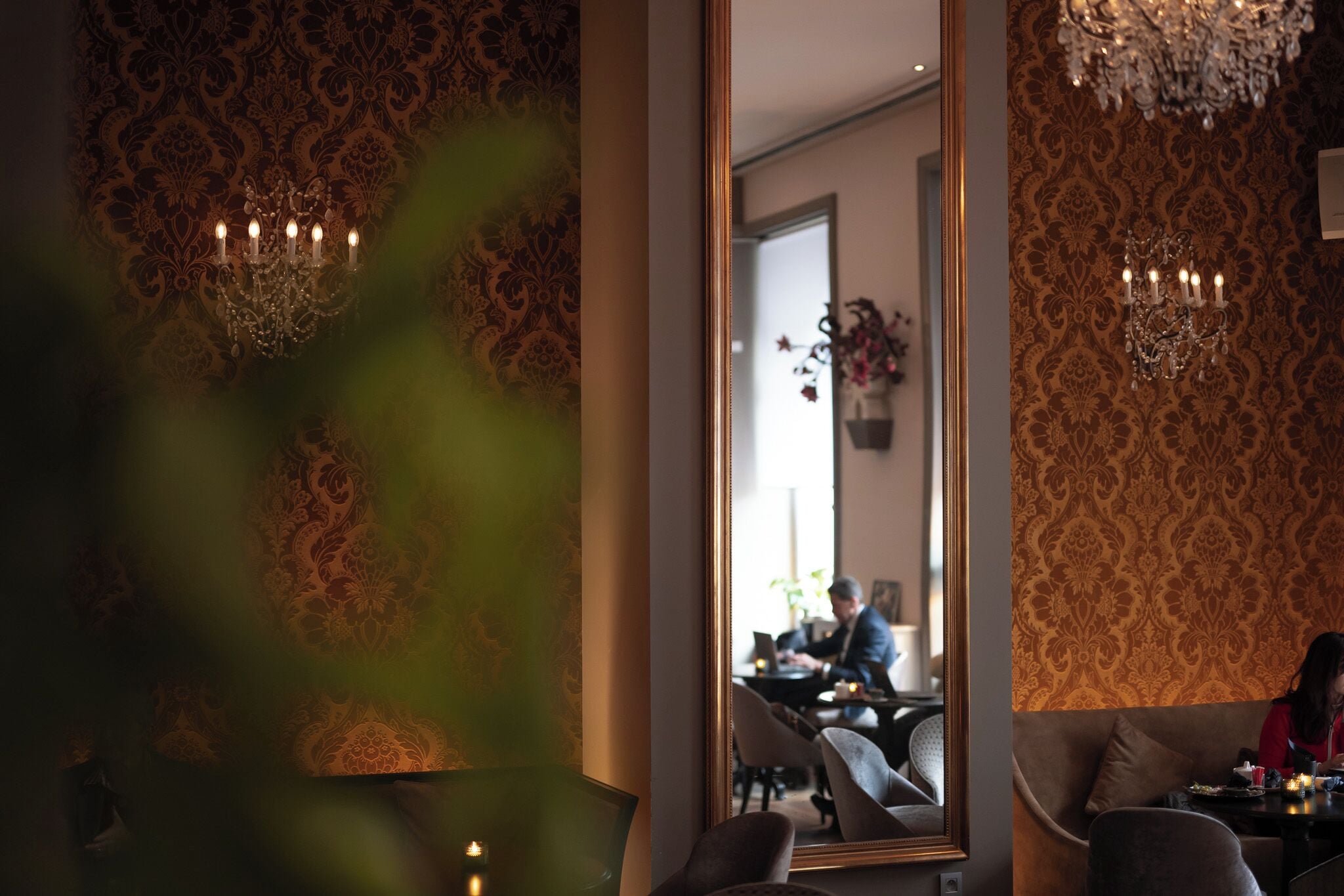 A room with patterned wallpaper, a chandelier, and a mirror reflecting a person working at a desk.