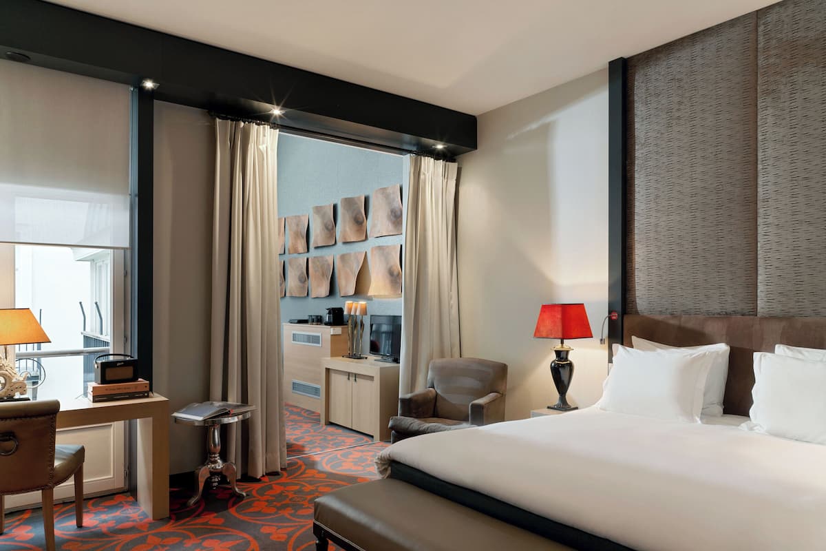 Une chambre d’hôtel avec un grand lit, un bureau avec une lampe, une chaise, une petite table avec une radio, une télévision et une armoire.