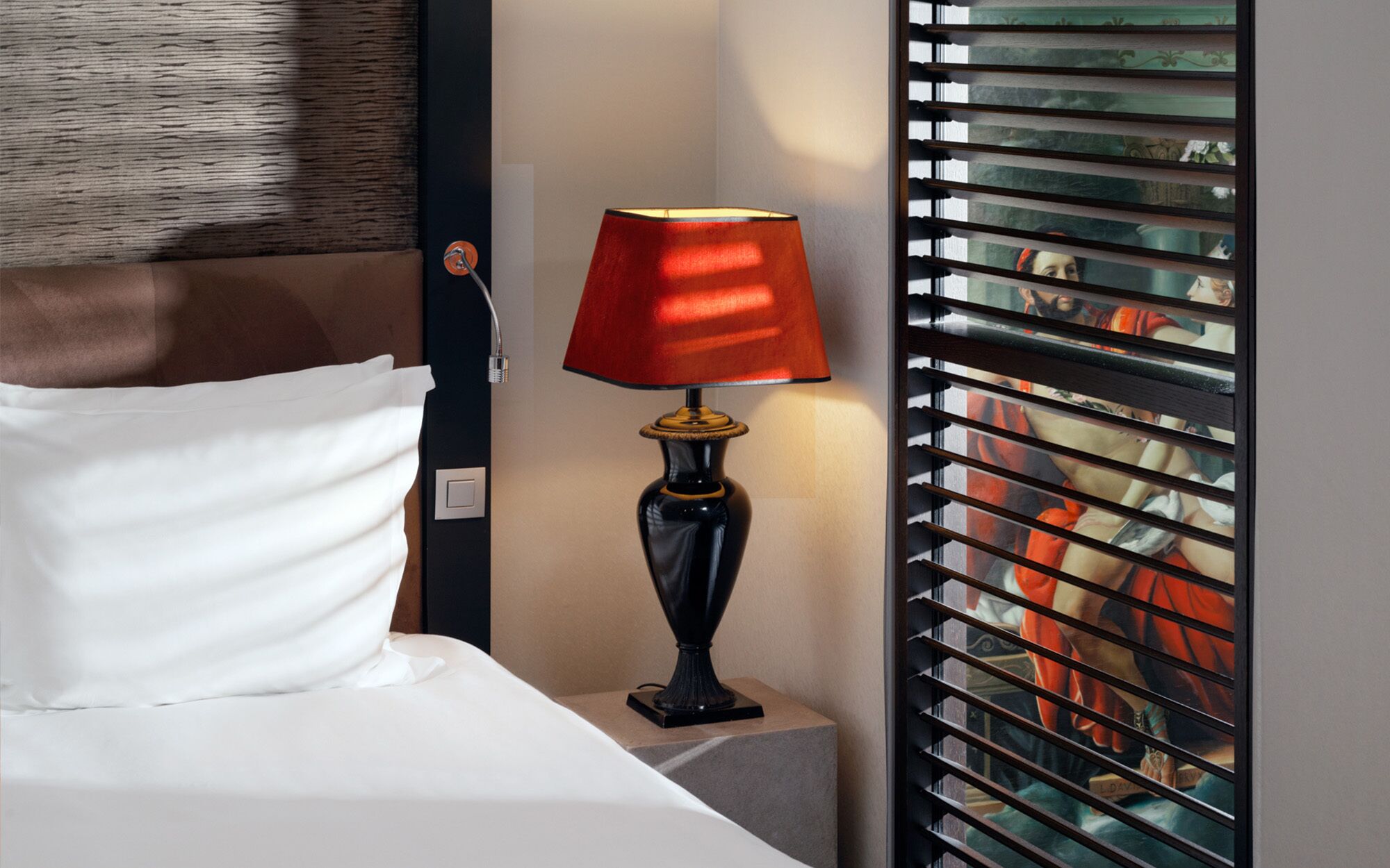 A hotel room with a bed, a red lamp, and a window with blinds.