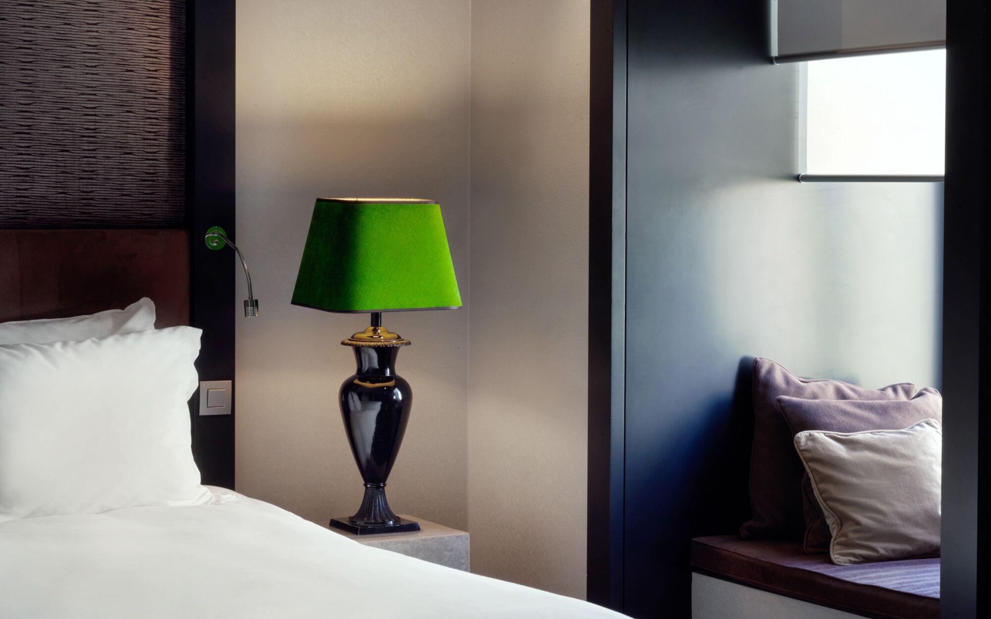 A neatly made bed with white linens, a green lamp on a bedside table, and a view of a window with a dark interior.