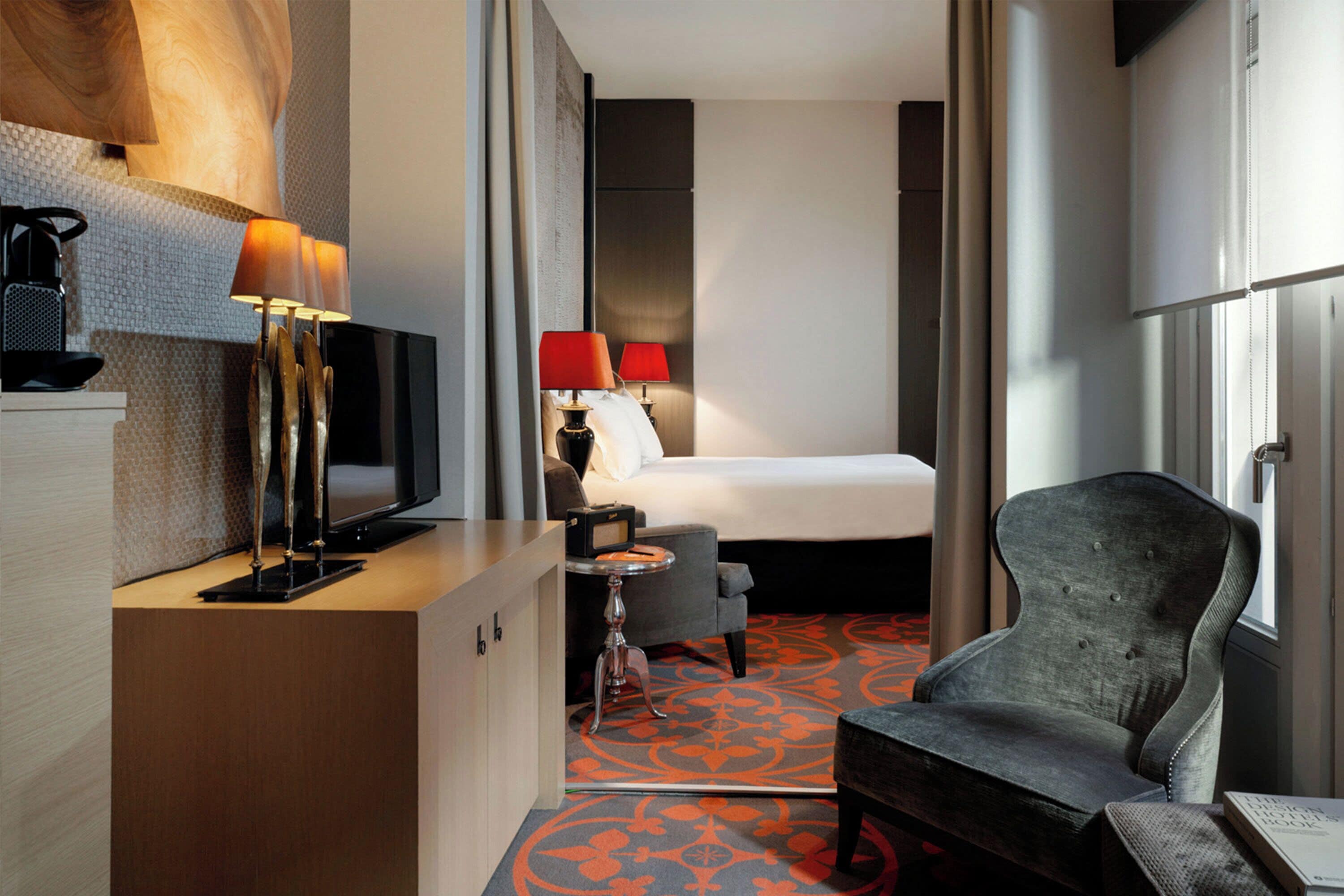 A hotel room with a bed, a desk with a television, two lamps, a chair, and a patterned carpet.
