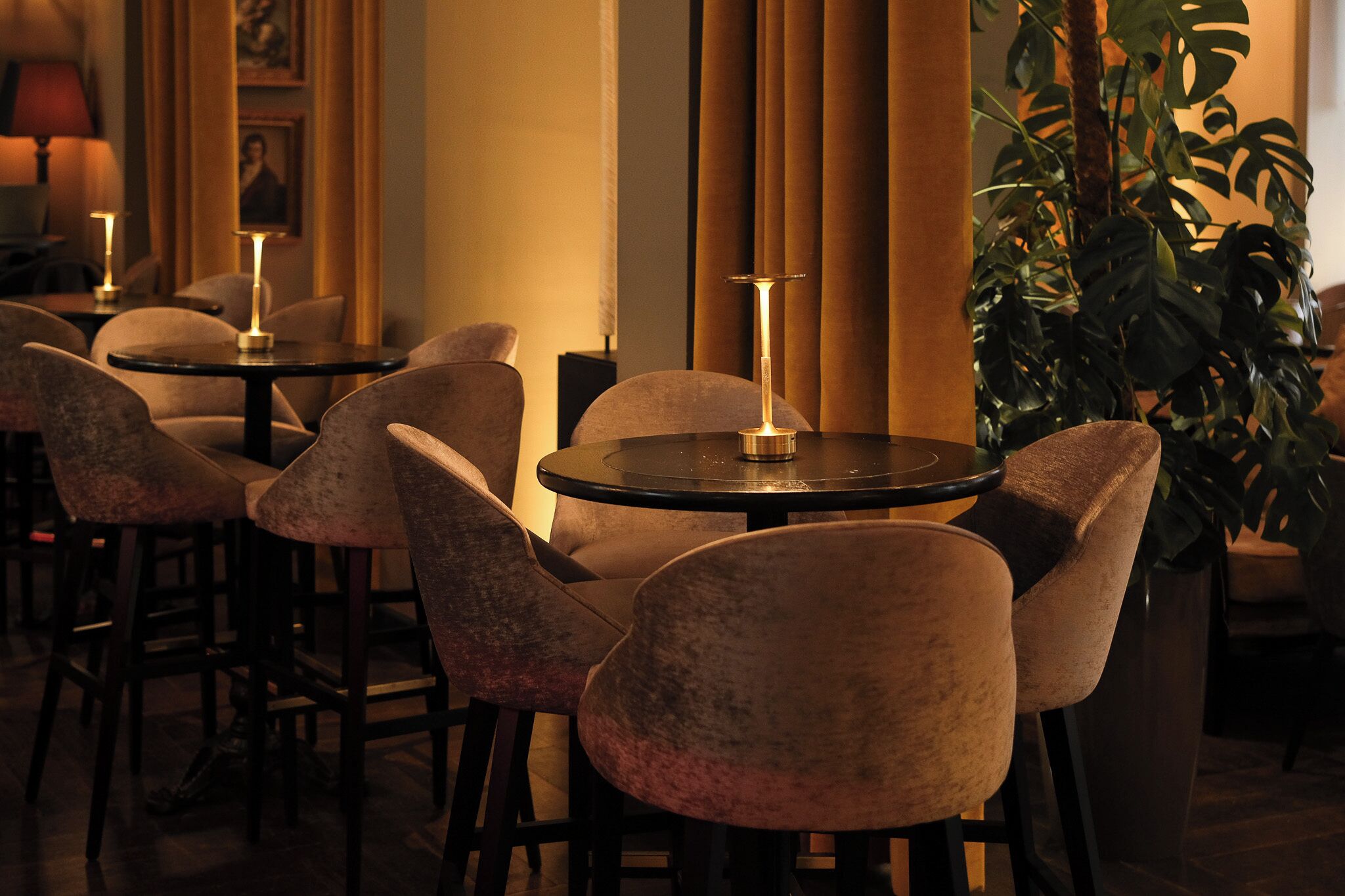 Un coin bar à la lumière tamisée, avec des tables rondes et des chaises rembourrées, une grande plante en pot et un portrait encadré accroché au mur.