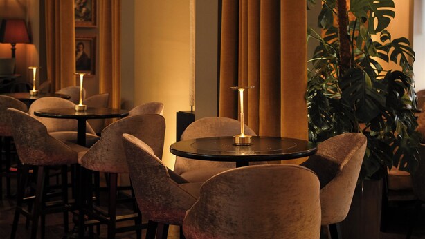 A dimly lit bar area with round tables and cushioned chairs, a large potted plant, and a framed portrait on the wall. - The Dominican, Brussels, a Member of Design Hotels (Brussels)