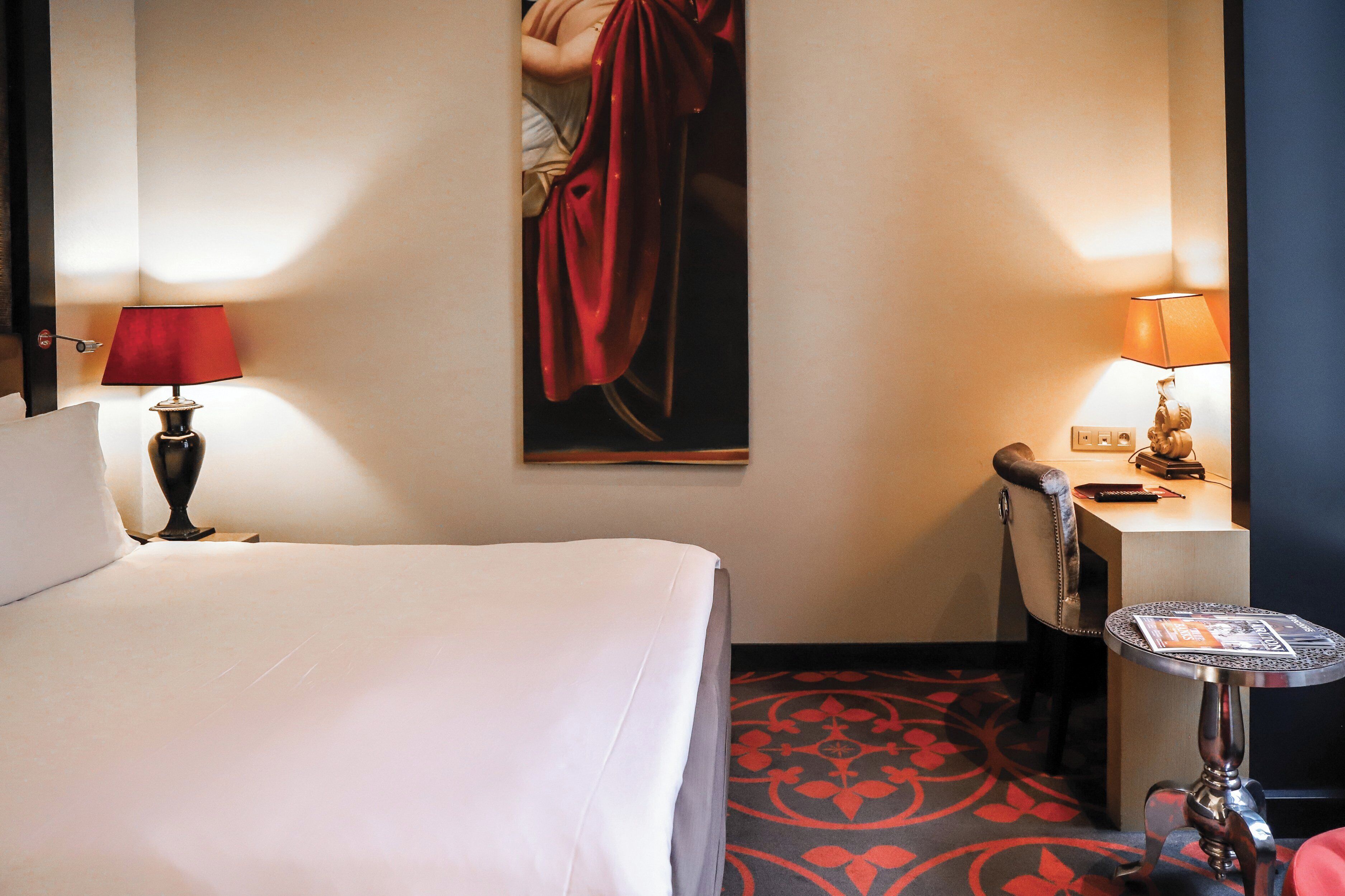 A hotel room with a bed, a desk, a chair, a small table, and a painting on the wall.