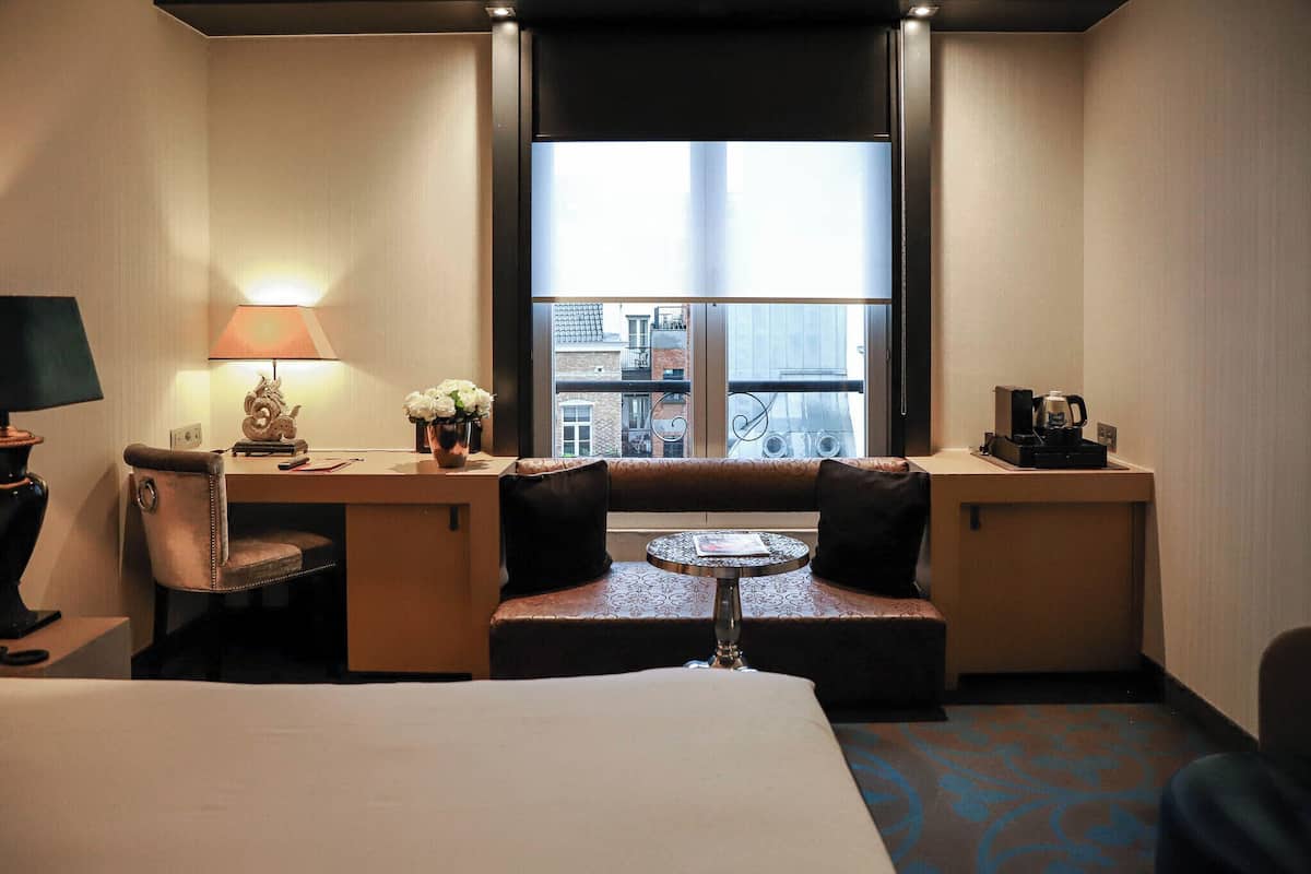 Une chambre d’hôtel comprenant un lit, un bureau avec une lampe, une petite table avec un vase de fleurs, un canapé donnant sur une fenêtre et une machine à café posée sur le bureau.