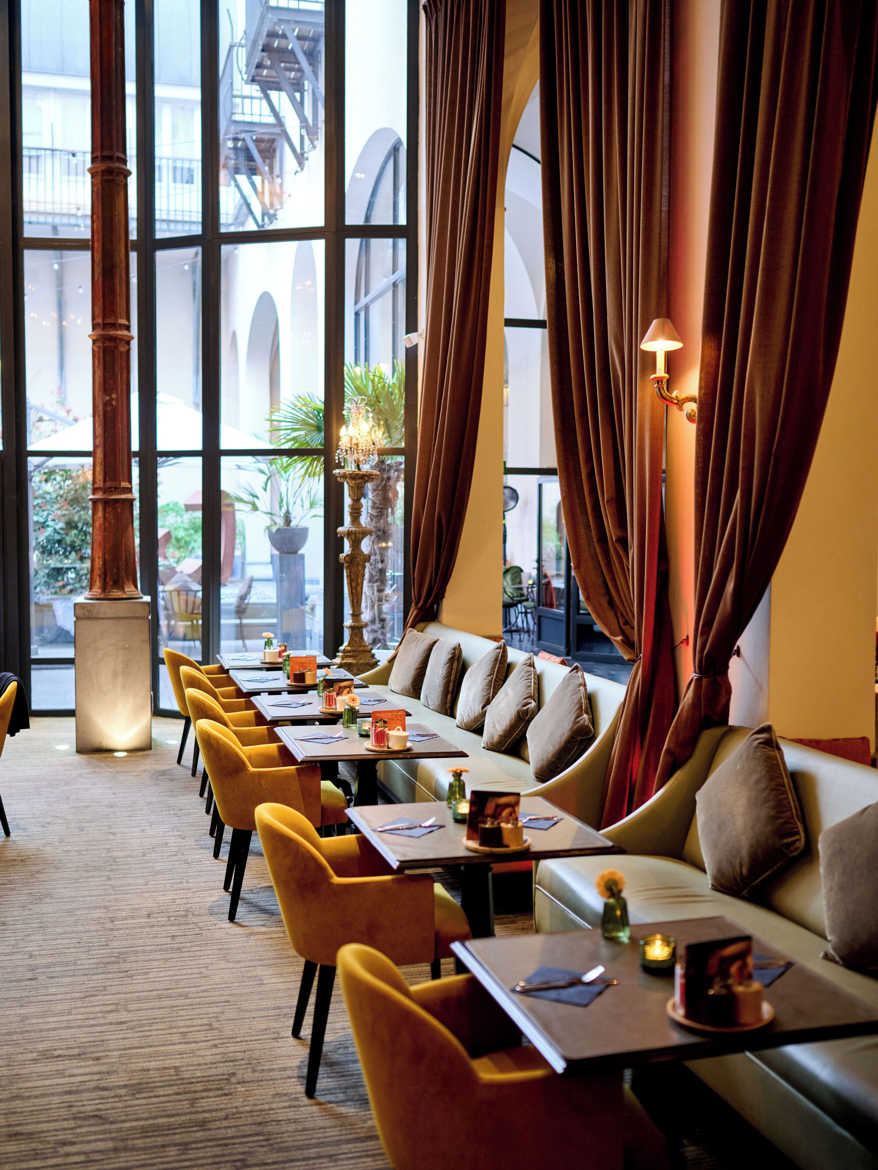 A hotel dining area with a long table, yellow chairs, a sofa, and large windows with curtains.
