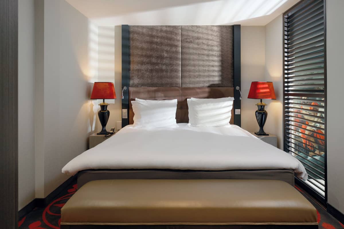 Une chambre d’hôtel avec un grand lit, deux lampes de chevet, un banc et une fenêtre avec des stores.