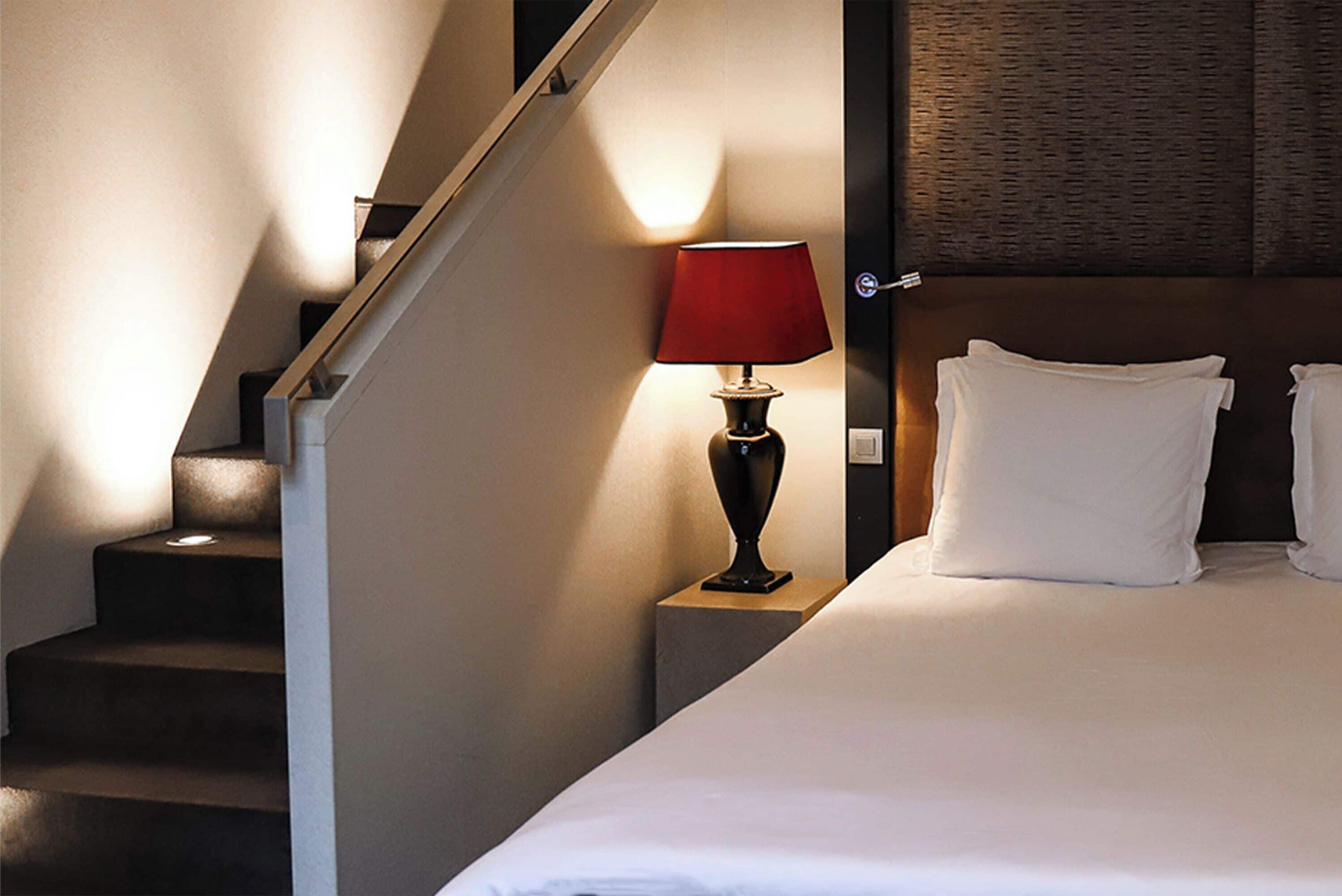 A hotel room with a bed, a nightstand with a lamp, and a staircase.