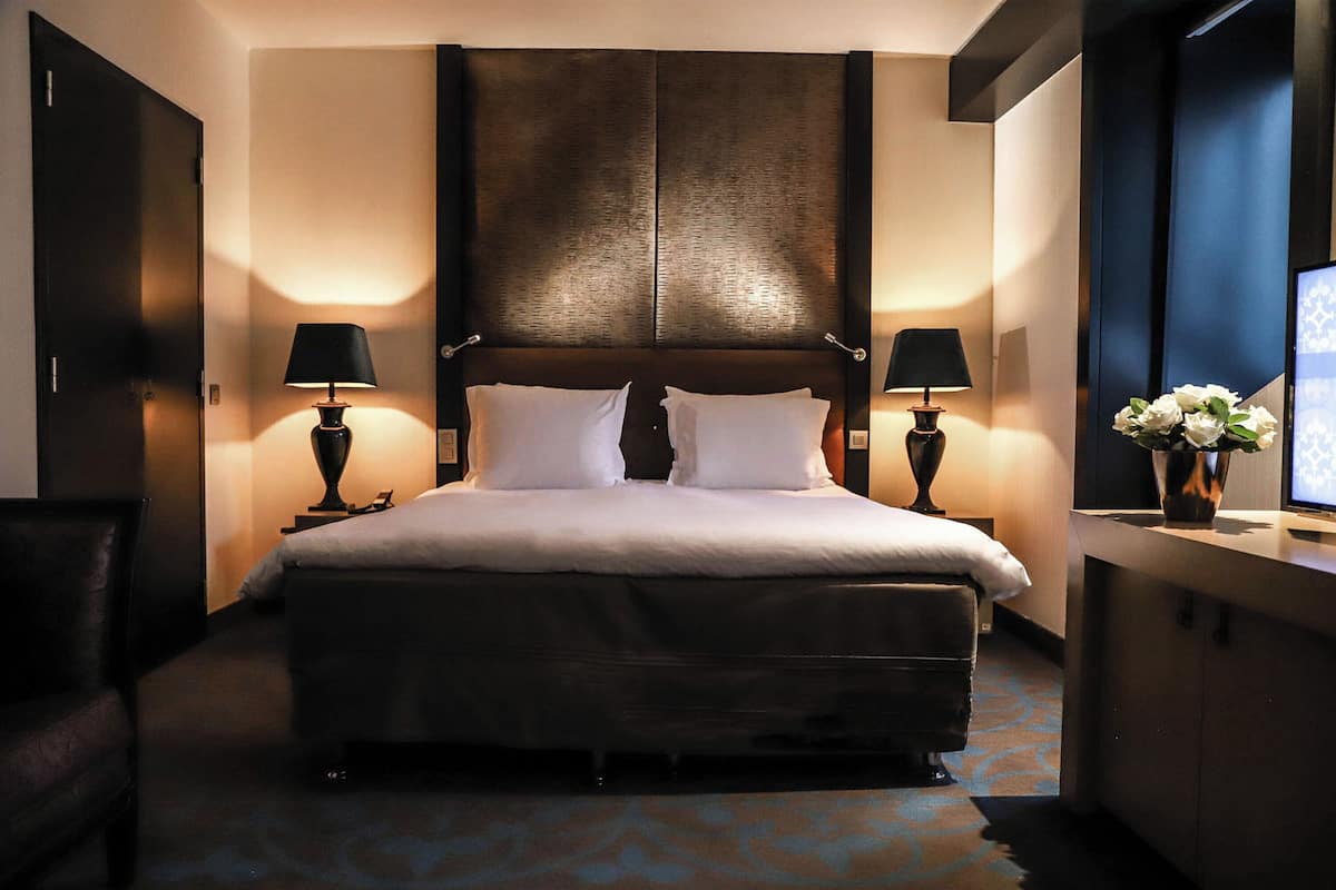 Une chambre d’hôtel avec un grand lit, deux tables de chevet avec des lampes, un bureau agrémenté d’un vase de fleurs et un téléviseur placé sur le mur de droite.