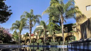 Piscina com reflexos nítidos, cercada por palmeiras e um edifício com arcos.