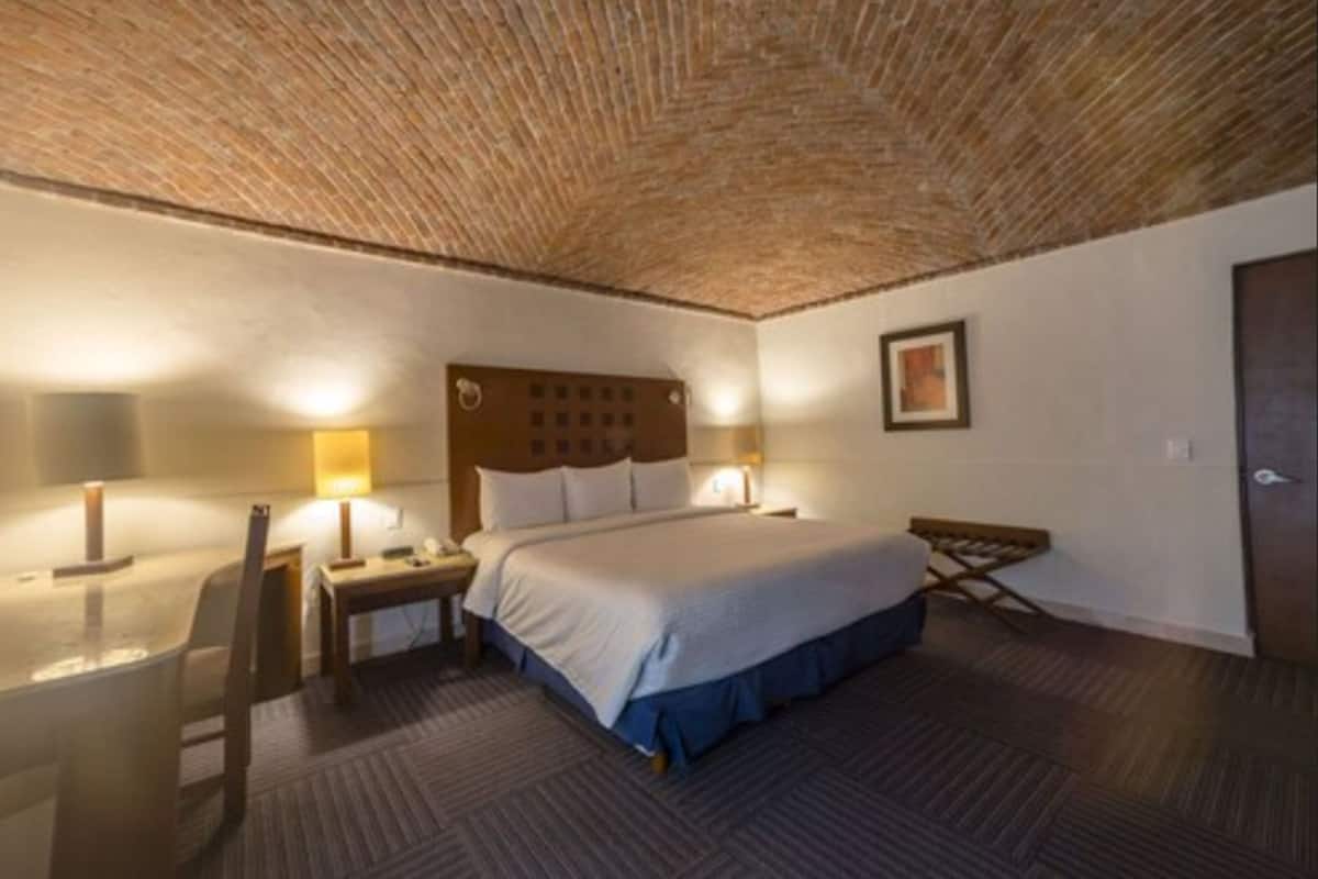 Ein Hotelzimmer mit einem großen Bett, einem hölzernen Kopfteil, zwei Nachttischen mit Lampen, einem kleinen Schreibtisch und einem Sessel.