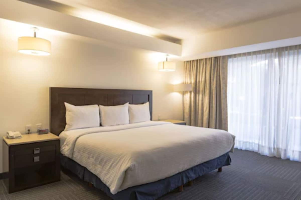 Ein Hotelzimmer mit einem großen Bett, zwei Nachttischlampen, einem Nachttisch und einem Fenster mit Vorhängen.