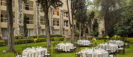 A courtyard with round tables set up for dining, surrounded by trees and a building with balconies.
