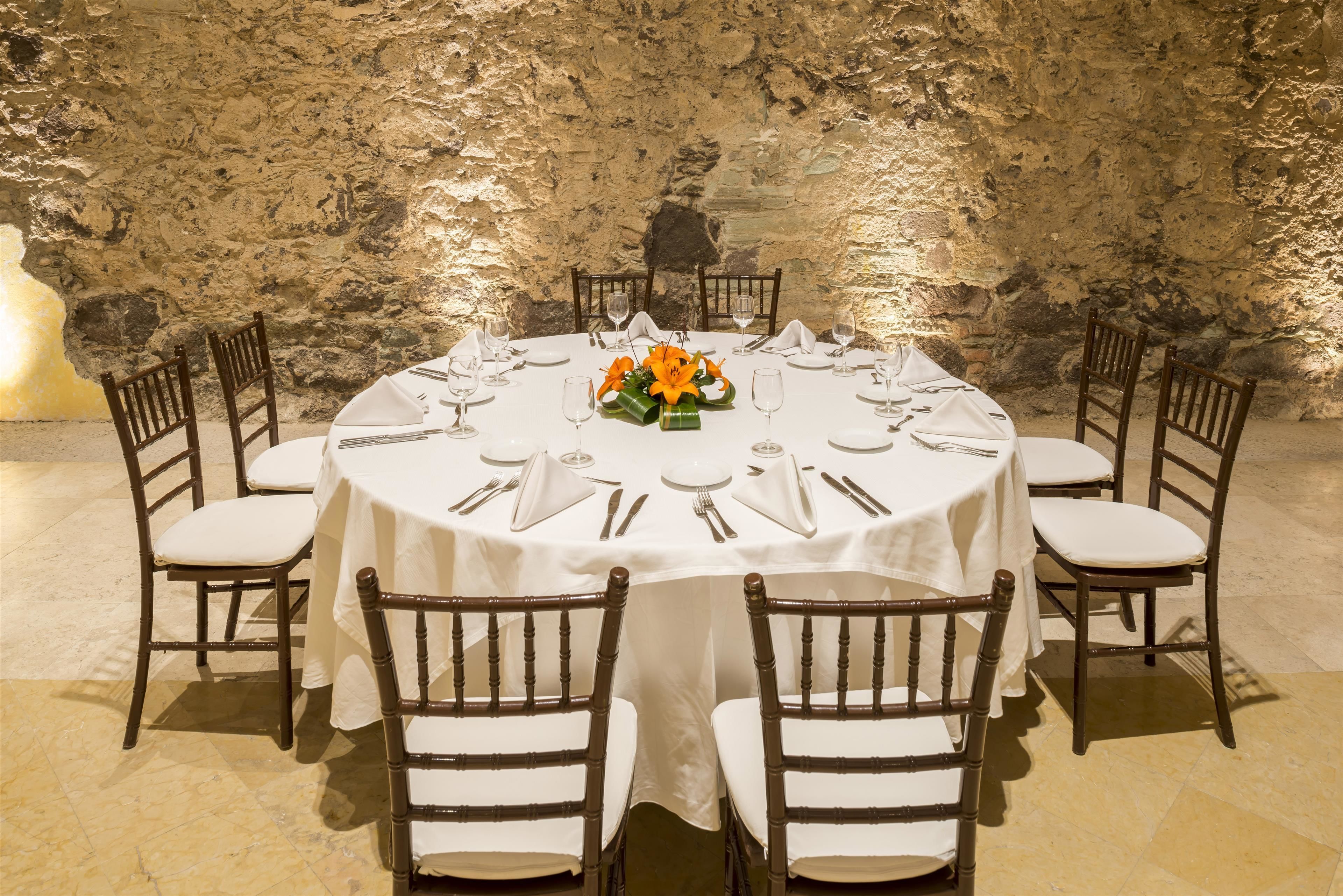 Une table ronde dressée pour huit personnes, recouverte de nappes blanches, avec des chaises en bois et un centre de table composé de fleurs oranges.