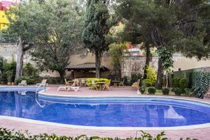 A swimming pool with lounge chairs and a table, surrounded by trees and a building with a tiled roof.
