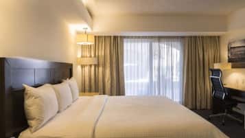 A hotel room with a large bed, a desk, and a chair. There is a window with curtains and a lamp on the desk.