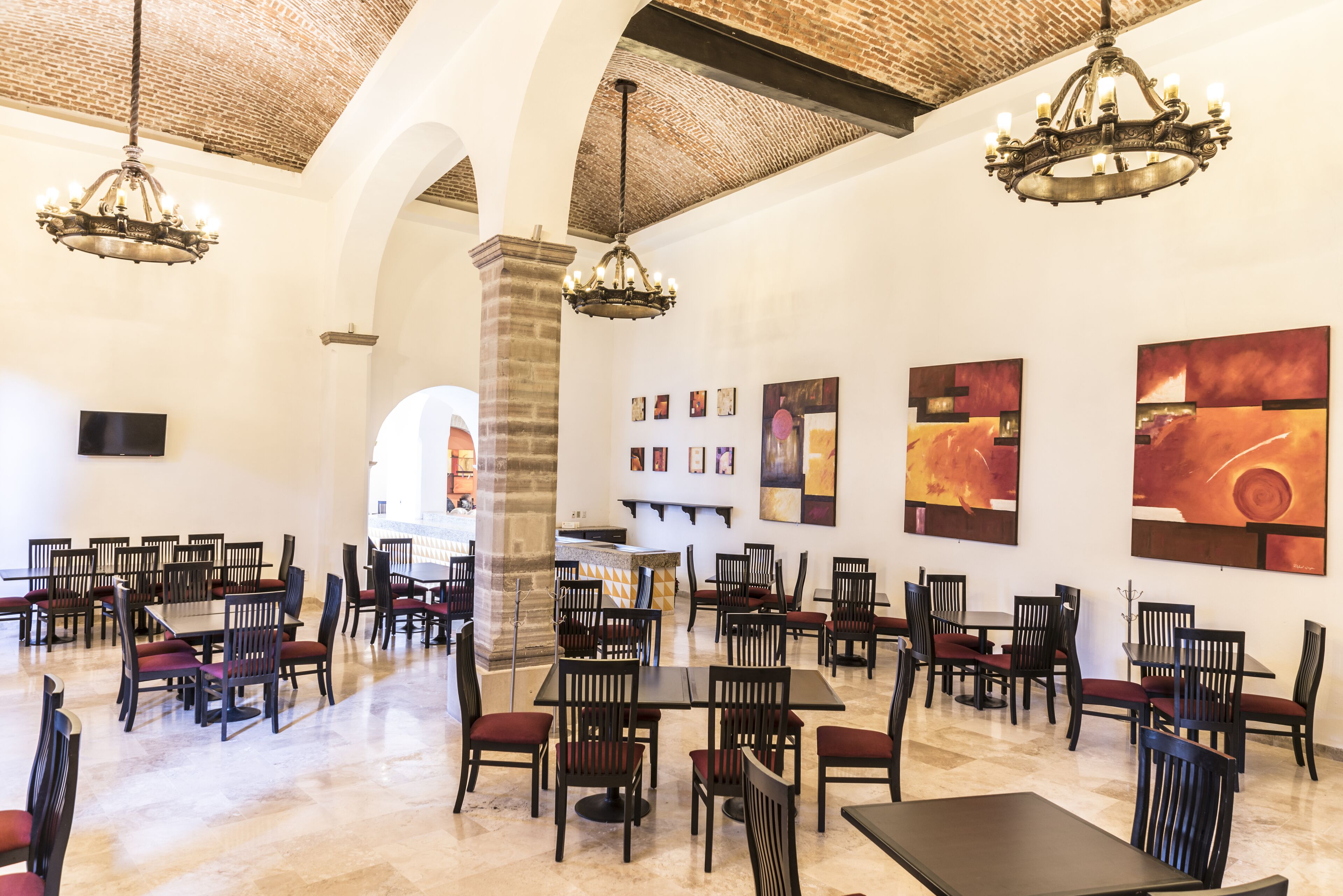 A spacious dining area with high ceilings, exposed brick, and modern chandeliers. Tables and chairs are arranged for dining.