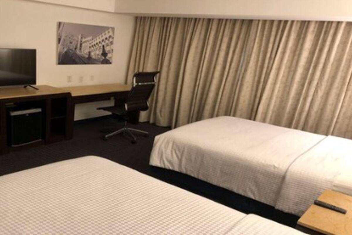 Ein Hotelzimmer mit zwei Betten, einem Schreibtisch mit Fernseher, einem Sessel und einem großen Fenster mit Vorhängen.