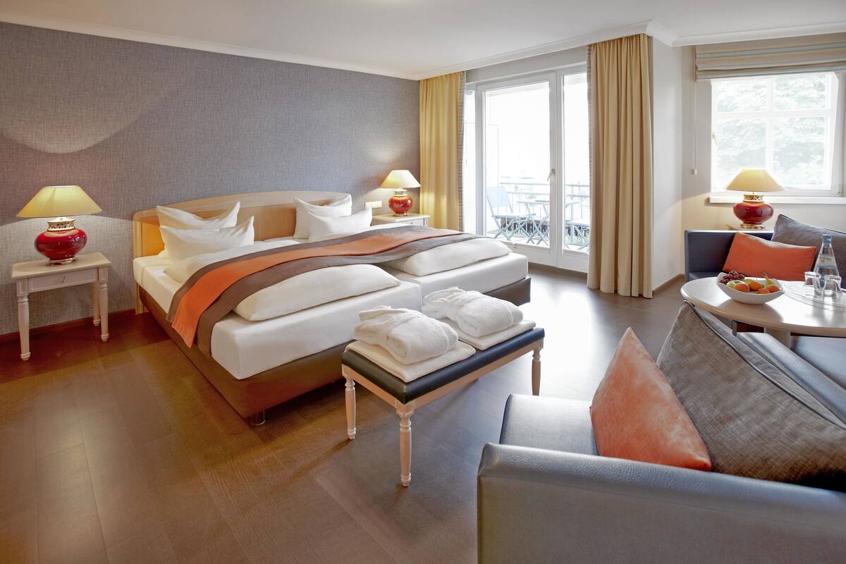 Ein Hotelzimmer mit Bett, Nachttischen, einer Bank, einem Sofa, einem Esstisch und einem Balkon.