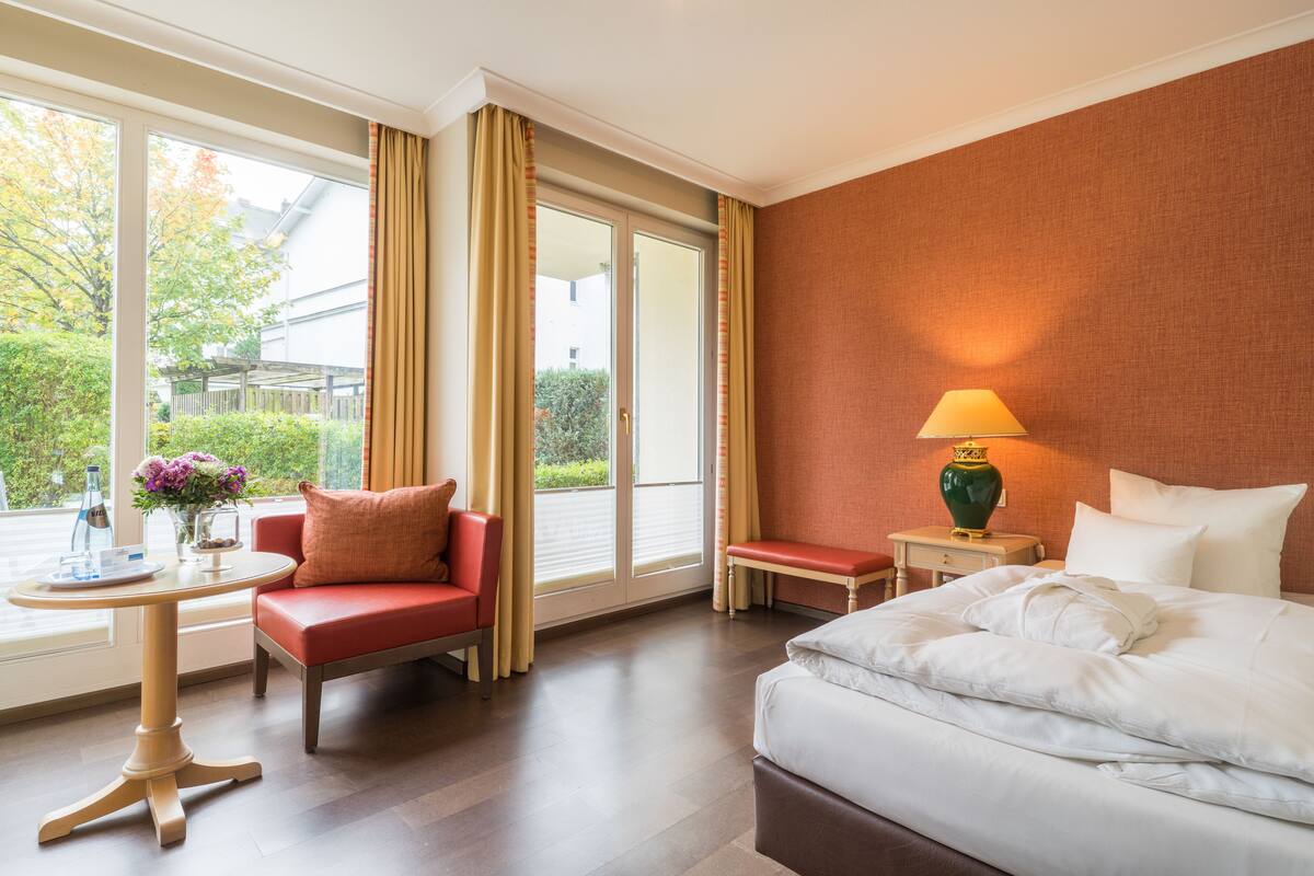 Ein Hotelzimmer mit einem Bett, einem Sessel, einem kleinen Tisch und einer Lampe. Ein Fenster mit Vorhängen bietet Blick auf Grünfläche.