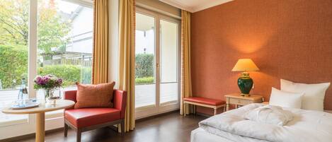 A hotel room with a bed, a chair, a small table, and a lamp. There is a window with curtains and a view of greenery outside.