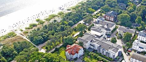 An aerial view of a coastal residential area with a large building complex, a beach with people, and dense greenery.