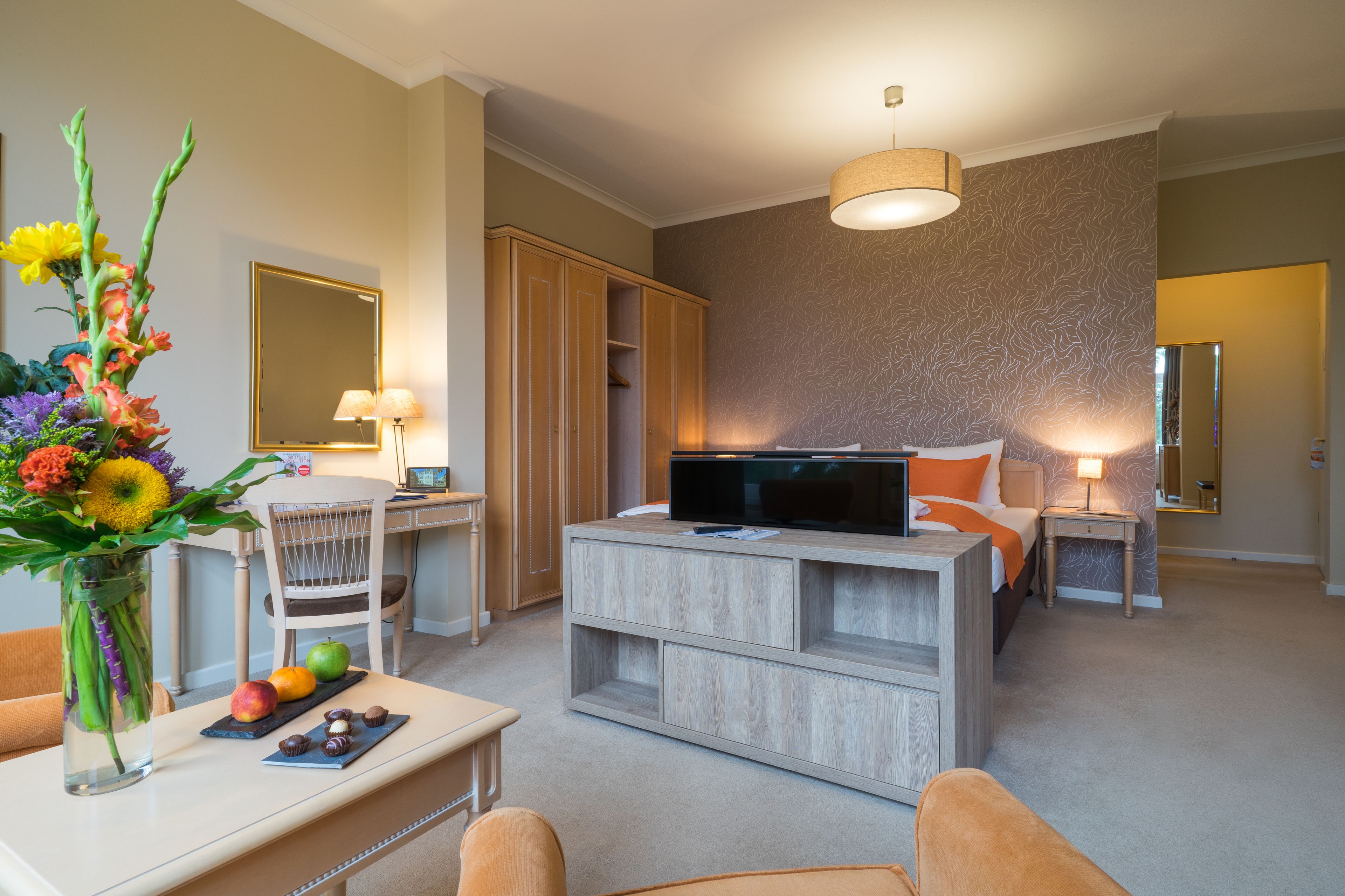 Una habitación de hotel moderna con un mueble de televisión de madera, un escritorio con una lámpara, una silla y un espejo grande.