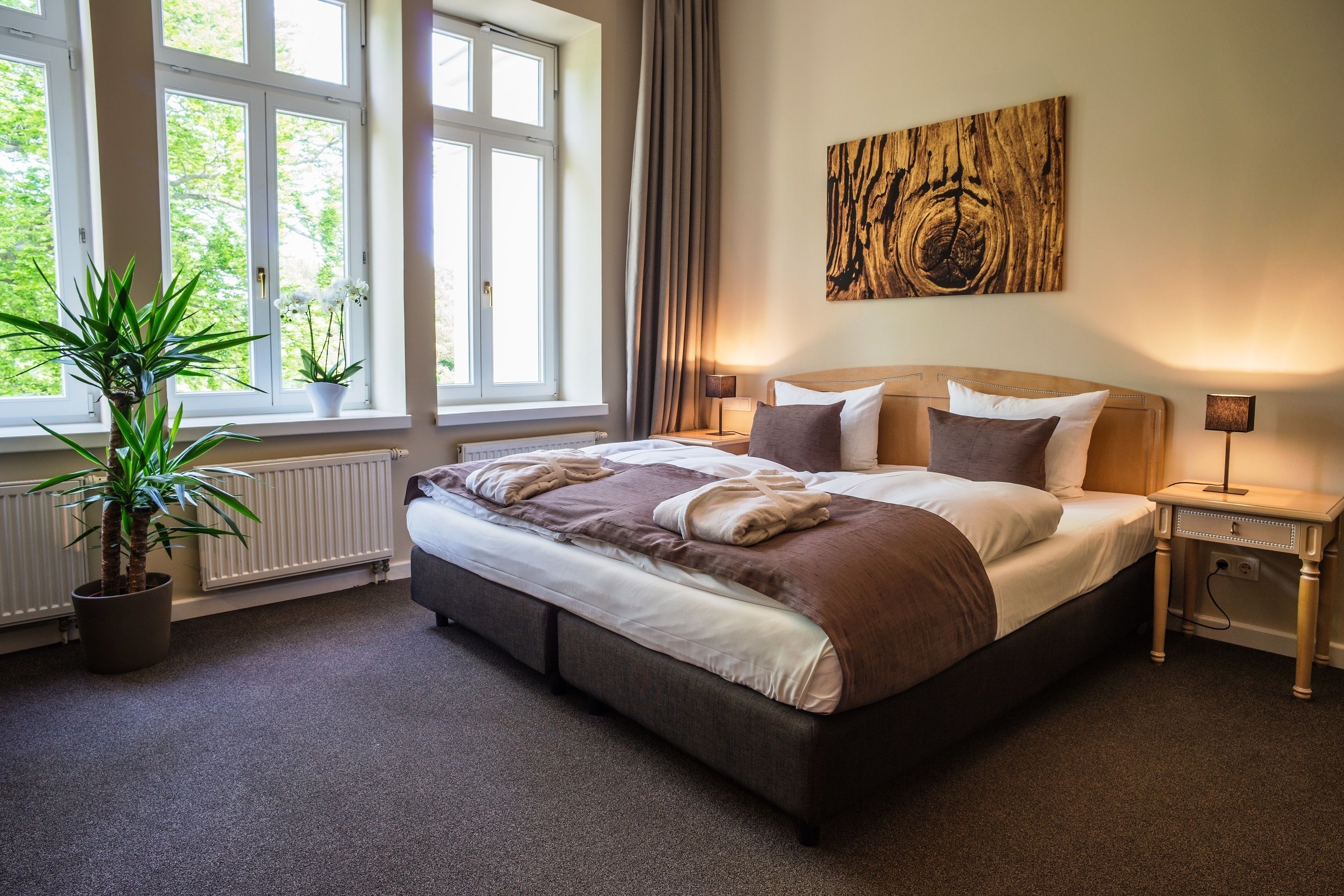 A neatly made bed with a brown and white color scheme, a large window with curtains, a potted plant, and a wooden wall art piece.