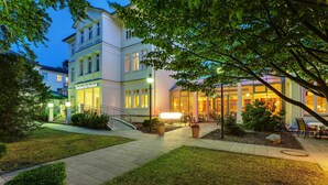 Hotel com entrada bem iluminada, área de estar coberta ao ar livre e jardim ajardinado.