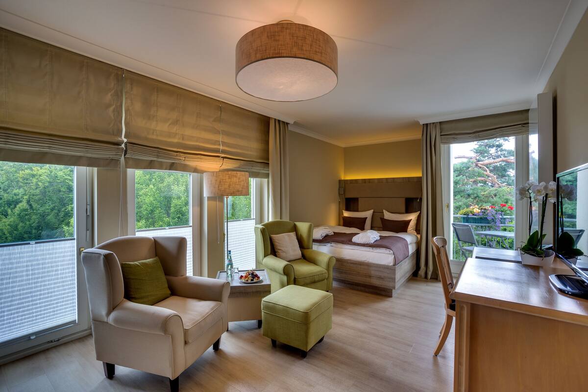 Ein modernes Hotelzimmer mit einem Bett, einem Schreibtisch, einem Sessel und einer Couch. Ein Balkon bietet Blick auf Bäume und Blumen.