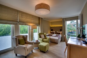 Une chambre d’hôtel moderne équipée d’un lit, d’un bureau, d’une chaise et d’un canapé. Elle dispose également d’un balcon avec vue sur les arbres et les fleurs.