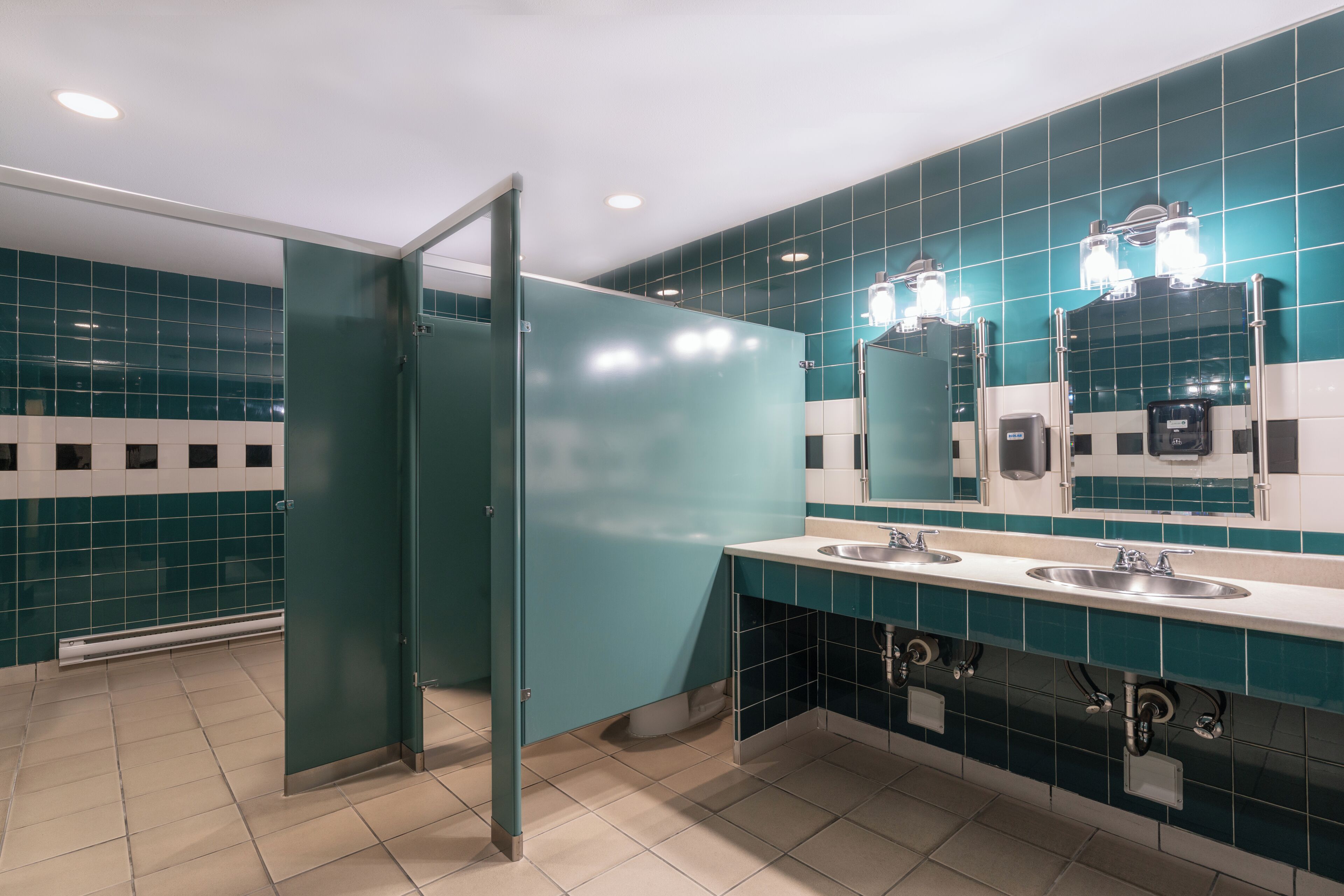 A restroom with teal tiles, two sinks, a mirror, and a glass partition.