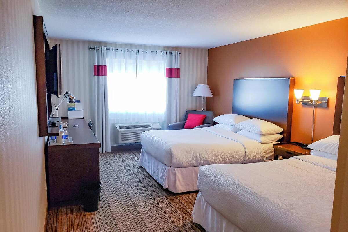 Ein Hotelzimmer mit zwei Betten, einem Schreibtisch mit Lampe, einem Fernseher und einem Fenster mit Vorhängen.