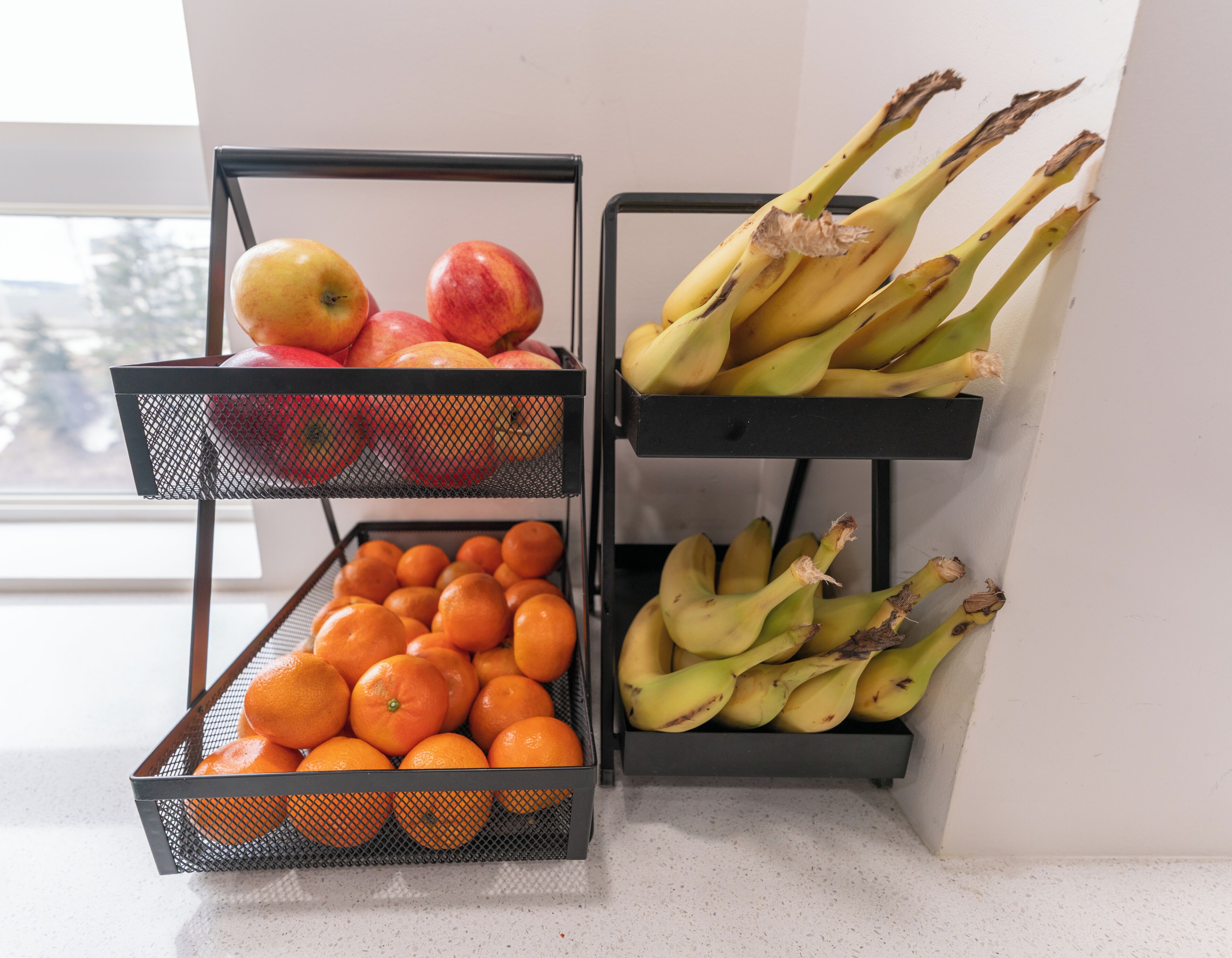 Two metal mesh fruit baskets on a countertop, one containing apples and oranges, the other bananas.