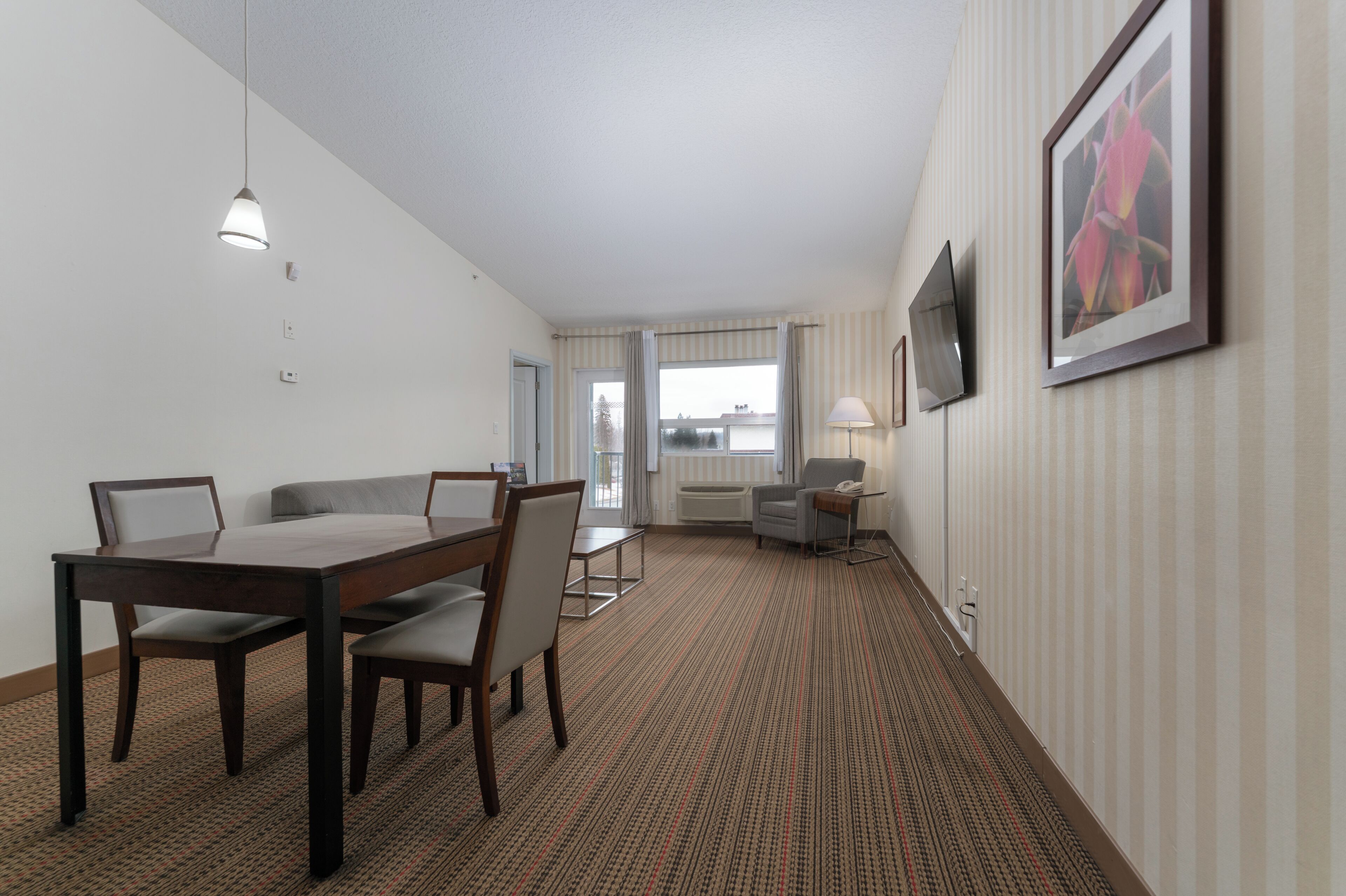 Habitación de hotel con una mesa de comedor, sillas, televisor, un cuadro enmarcado y una ventana con cortinas.