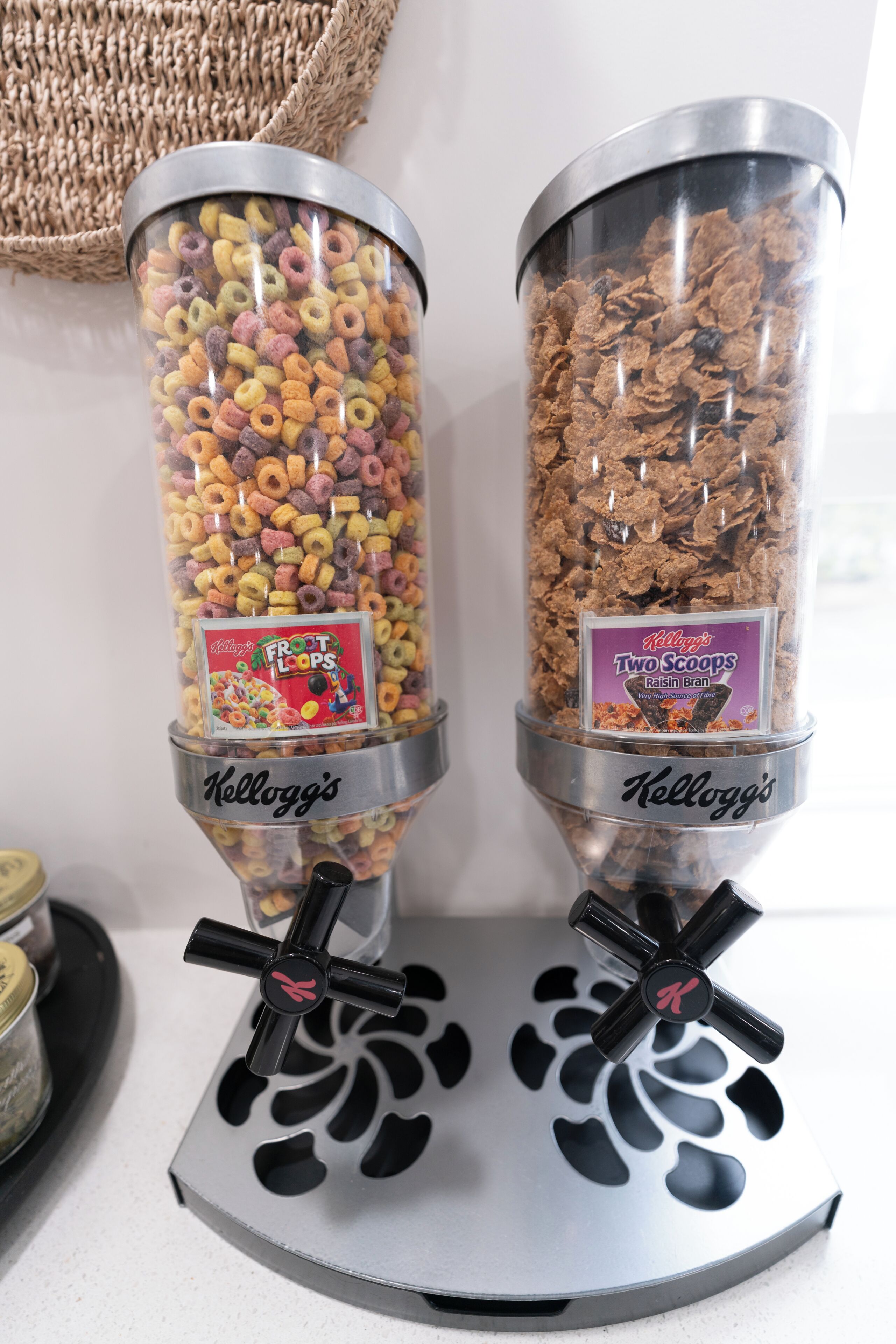 Two cylindrical dispensers filled with cereal, labeled 'Kellogg's Frost Loops' and 'Kellogg's Two Scoops Raisin Bran,' placed on a metallic stand.