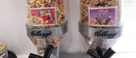 Dos dispensadores cilíndricos llenos de cereal, con la etiqueta 'Kellogg's Frost Loops' y 'Kellogg's Two Scoops Raisin Bran', ubicados sobre un soporte metálico.